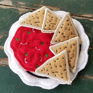 May include: A white plate with a scalloped edge, decorated with a red felt salsa dip and four brown felt tortilla chips.