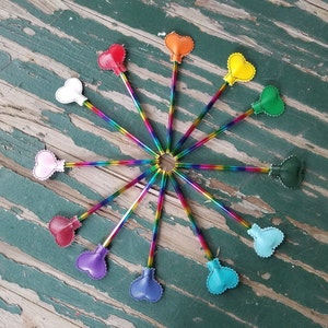 Könnte beinhalten: Ein Set mit 12 regenbogenfarbenen Bleistiften mit herzförmigen Aufsätzen in verschiedenen Farben, darunter rosa, rot, orange, gelb, grün, blau, lila und weiß. Die Bleistifte sind in einem Kreis auf einer Holzoberfläche angeordnet.