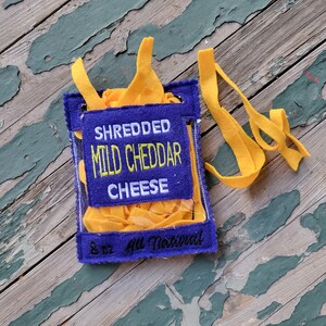 May include: A purple felt bag with a clear plastic window, filled with yellow felt strips. The bag has the text "Shredded Mild Cheddar Cheese 8 oz All Natural" embroidered on it.