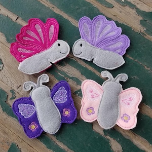 May include: Four felt butterfly appliques in various colors. The butterflies are pink, purple, gray, and pink with purple accents. The butterflies have embroidered details and are perfect for crafting projects.