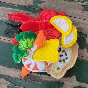 May include: A felt food set with a red lobster, green broccoli, yellow lemon wedges, a yellow and white bread slice, a white shrimp, an orange carrot, and a brown bread slice.