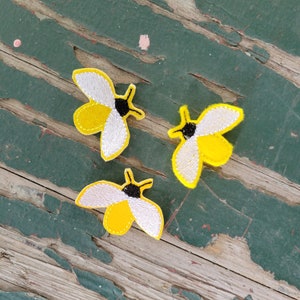 May include: Three yellow and white felt bee patches with black eyes and wings. The patches are on a green wooden surface.
