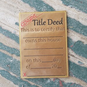 May include: A gold-colored card with the text "GENUINE Title Deed" and "This is to certify that ____ owns this house on this ____ day of ____ 20____" with an "X" at the bottom.
