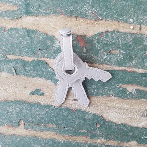 May include: A silver leather keychain with two key shapes. The keychain is hanging from a weathered wooden surface.
