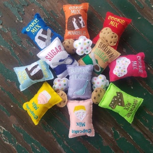 May include: A collection of felt play food bags, including cake mix, cookie mix, brownie mix, cupcake mix, and baking flour. The bags are decorated with colorful embroidery and include small felt cookies and other food items.