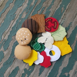 May include: Felt food play set with a hamburger, bun, lettuce, tomato, cheese, and pickle. The set is made of felt and is perfect for pretend play.