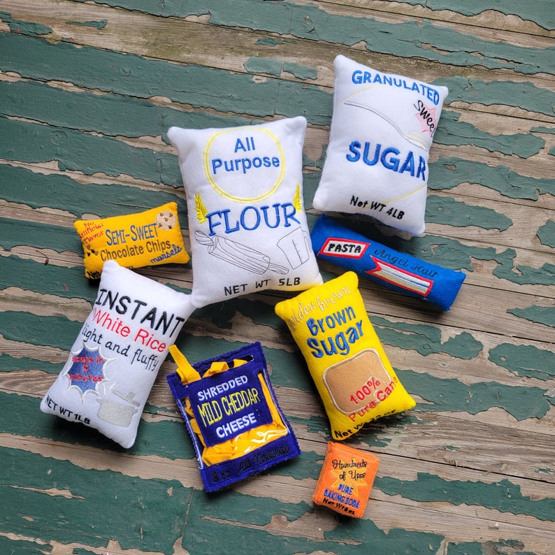 May include: A collection of eight yellow and white fabric pouches with embroidered text, including "All Purpose Flour", "Granulated Sugar", "Brown Sugar", "Instant White Rice", "Shredded Mild Cheddar Cheese", "Semi-Sweet Chocolate Chips", "Pasta Angel Hair", and "Pure Baking Soda".