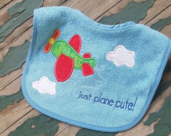 May include: A light blue baby bib with an embroidered airplane and clouds. The bib has the text "just plane cute!" embroidered on it.