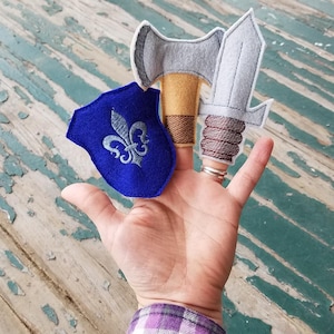 May include: A set of three felt finger puppets depicting a shield with a fleur-de-lis, an axe, and a sword. The puppets are gray, brown, and blue.