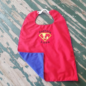 Peut inclure: Cape de super-héros rouge et bleue avec un logo Superman jaune et le texte "Captain Leon" brodé sur le devant.