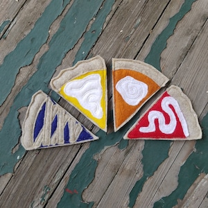 May include: Five felt pie slice ornaments in various colors: yellow, orange, red, and blue. Each slice has a tan crust and white embroidered details. The ornaments are arranged on a weathered wooden surface.