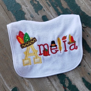 Könnte beinhalten: Weißer Lätzchen mit dem gestickten Namen "Amelia" in rotem Garn. Das "A" ist mit einem Pilgerhut und Federn verziert. Ein kleiner Kürbis ist zwischen dem "m" und dem "e" gestickt.