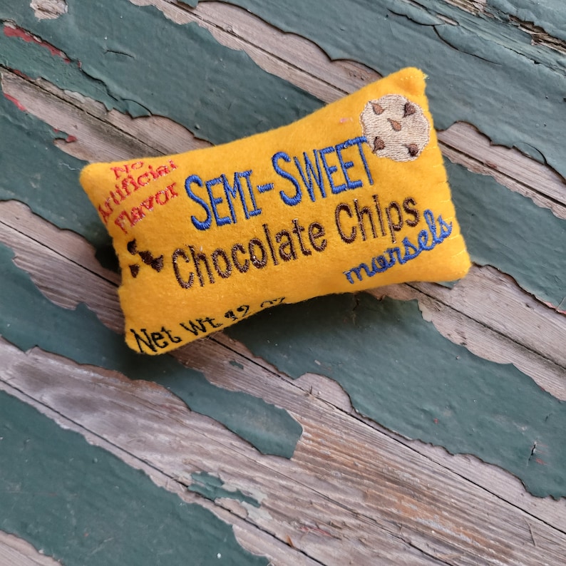 May include: A yellow fabric pillow shaped like a chocolate bar with the text "No Artificial Flavor Semi-Sweet Chocolate Chips morsels Net Wt 1.9 oz" embroidered on it.