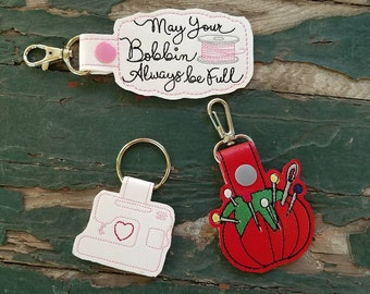 May include: Three sewing-themed keychains. One keychain is a white sewing machine with a red heart. Another keychain is a red pincushion with a variety of colorful pins. The third keychain is a white tag with the text "May Your Bobbin Always Be Full" and a pink spool of thread.