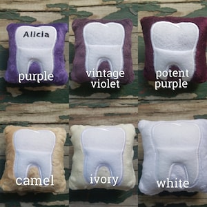 May include: Six plush tooth pillows in different colours: purple, vintage violet, potent purple, camel, ivory, and white. Each pillow has a white embroidered tooth on the front. The pillows are arranged in two rows of three.