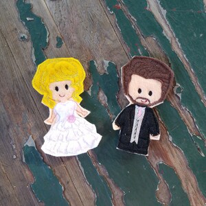 May include: A pair of felt finger puppets depicting a bride and groom. The bride is wearing a white dress with pink flowers and a yellow veil. The groom is wearing a black suit with a white shirt and a black bow tie.