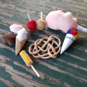 May include: A collection of felt food items, including a pink cotton candy, a red apple, a brown pretzel, a yellow hot dog, a brown and white ice cream cone, a blue and white snow cone, and two brown caramel apples.