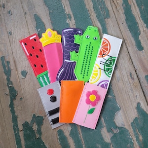Könnte beinhalten: Ein Set aus zehn bunten Stoff-Lesezeichen mit verschiedenen Designs, darunter eine Wassermelone, eine Krone, ein Blumenmuster, ein Zitrusfrüchte-Muster und ein Alligator. Die Lesezeichen sind alle etwa 25 cm lang.