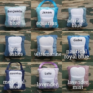 May include: Nine plush tooth-shaped cushions in various colours, including light blue, blue, ultramarine, royal blue, lavender, and lavender mist. Each cushion has a different name embroidered on it, such as Benjamin, Jaxon, Gabe, Houston, and Lulu.