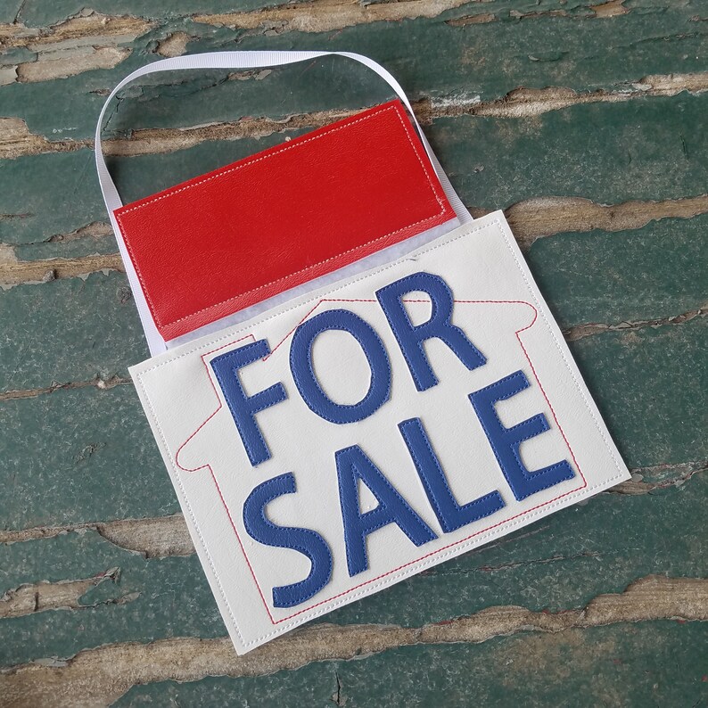 May include: White felt house-shaped sign with blue embroidered text that reads "FOR SALE". The sign has a red felt pocket on the back for hanging.