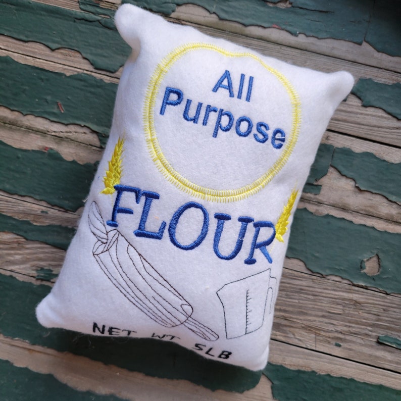 May include: White pillow shaped like a flour bag with blue embroidery that says "All Purpose Flour" and a yellow circle around the words. The pillow also has a drawing of a rolling pin and a measuring cup.