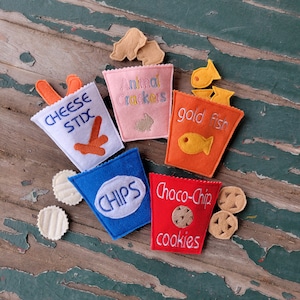 May include: Five felt pouches with different snack names and images. The pouches are: Cheese Sticks, Animal Crackers, Gold Fish, Chips, and Choco-Chip Cookies.