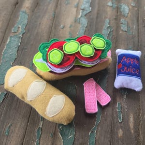Könnte beinhalten: Ein Filz-Spielzeug-Lebensmittelset bestehend aus einem beigen Baguette, einem Sandwich mit roten Tomaten, grünem Salat und Gurkenscheiben, zwei rosa Streifen sowie einer weißen Saftpackung mit blauem Aufdruck Apple Juice und einem Apfel.