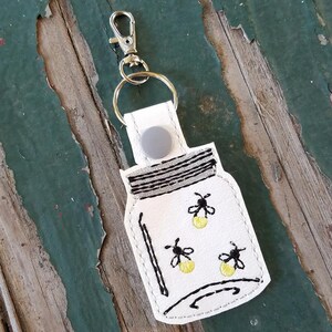 May include: White keychain featuring a mason jar design with three fireflies. The jar has black outlines and a silver clasp. The fireflies are yellow with black details. The keychain is attached to a silver metal clasp.