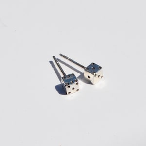 Könnte beinhalten: Ein Paar silberfarbene Würfel-Ohrstecker. Jeder Ohrring hat einen kleinen, quadratischen Würfel mit schwarzen Punkten, die Zahlen darstellen. Die Ohrringe haben einen Stecker für durchbohrte Ohren. Die Würfel sind vor weißem Hintergrund.