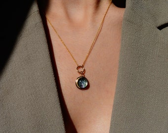 Könnte beinhalten: Eine goldene Kettenkette mit einem runden Anhänger mit einem blauen und grünen Edelstein. Der Anhänger ist in einen goldenen Rahmen gefasst.