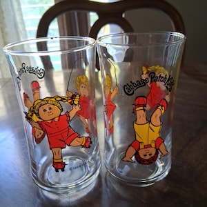 May include: Two clear glass tumblers featuring Cabbage Patch Kids graphics. One glass shows a character jumping rope, while the other shows a character doing a handstand. Both glasses include the Cabbage Patch Kids logo and 1984 OAA text.