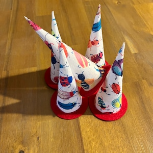 May include: Five white fabric cone-shaped party hats with red trim. Each hat is decorated with a colorful bug pattern.