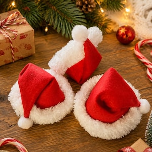 May include: Three miniature Santa hats made of red felt with white fuzzy trim and a small white pom-pom at the tip of each hat. The hats are arranged on a wooden surface, each featuring a conical shape with a soft, textured white border.