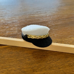 May include: A miniature white sailor hat with a black brim and a gold-colored braided band. The hat features a small black button on the side. The hat is placed on a wooden stick, showcasing its small size and craftsmanship.