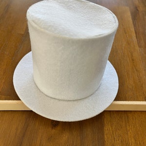 May include: A white felt top hat with a wide brim. The hat is sitting on a wooden surface.