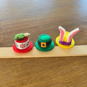 May include: Three miniature decorative hats: a red hat with a green light bulb, a green hat with a gold buckle, and a yellow hat with bunny ears. The hats are made of felt and are arranged on a wooden stick.