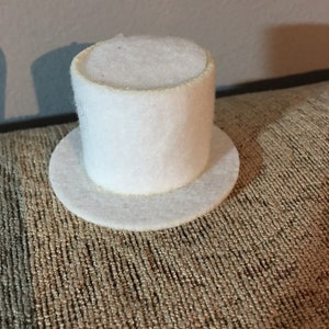 May include: A white felt top hat with a flat brim. The hat is small and would be suitable for a doll or miniature figure.