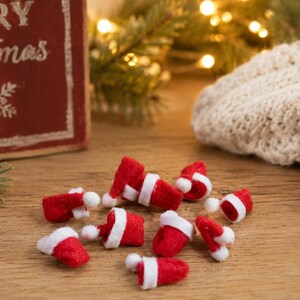 May include: A collection of small, miniature Santa Claus hats made of red felt with white trim and a white pom-pom at the tip. These tiny festive accessories are scattered across the surface, each featuring a classic conical shape and white band.