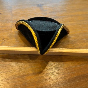 May include: A miniature black tricorn hat with gold trim. The hat is made of a felt-like material and has a triangular shape. The gold trim outlines the edges of the hat, adding a decorative touch. The hat is likely a decorative item or a costume accessory.