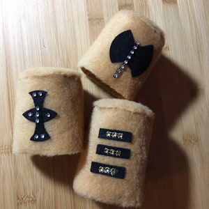 May include: Four tan fuzzy wrist cuffs with black felt designs and rhinestones. The designs include a geometric shape, a diamond, and three horizontal lines.