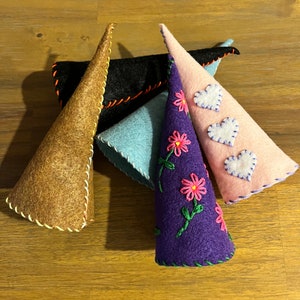 May include: Four felt cone hats with different colors and embroidery. One hat is brown with yellow stitching, one is black with red stitching, one is light blue with black stitching, and one is purple with pink flowers and green leaves.