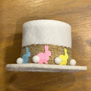 May include: A white felt Easter hat with a brown burlap band decorated with colorful bunny silhouettes and white pom-poms.