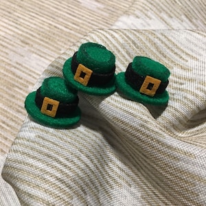 May include: Three green felt hats with black bands and gold buckles. The hats are arranged in a row on a white and brown patterned fabric.