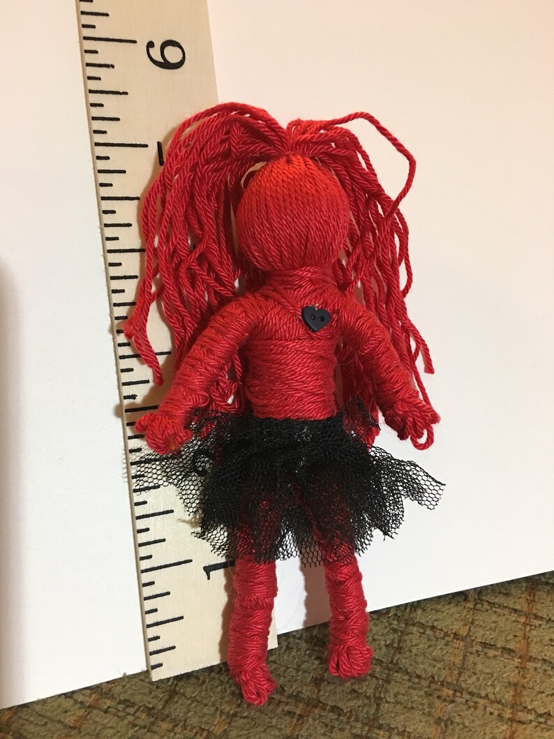 May include: A red yarn doll with long red yarn hair, wearing a black tulle skirt and a black heart-shaped button. The doll is standing next to a wooden ruler.