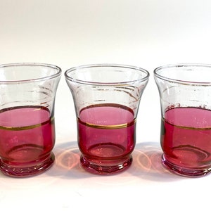 May include: Three clear glass drinking glasses with a pink base and a gold band around the middle.