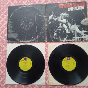 May include: A double vinyl record set titled Grand Funk Live Album. The gatefold cover features a black and white concert photograph. Two black vinyl discs are included, each with a bright yellow center label featuring the Capitol Records logo.