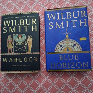 May include: Two hardcover books by Wilbur Smith. The black book is titled Warlock: A Novel of Ancient Egypt and features Egyptian figures and a winged sun disk. The blue book is titled Blue Horizon and features a large, detailed compass rose design.