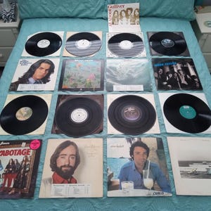 May include: A collection of vinyl records, including albums by various artists. The records are black with visible labels and covers featuring portraits and band photos. The albums are arranged on a blue bedspread.