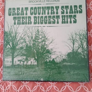 May include: A vintage vinyl record album titled "Great Country Stars Their Biggest Hits" by Brookville Records. The album cover features green text and a black and white landscape scene of a house and trees.