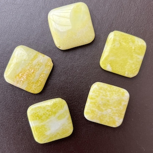 May include: Five square-shaped gemstones with rounded corners. The stones are primarily yellow with white marbling and a glossy finish. The gemstones are arranged on a dark brown surface.